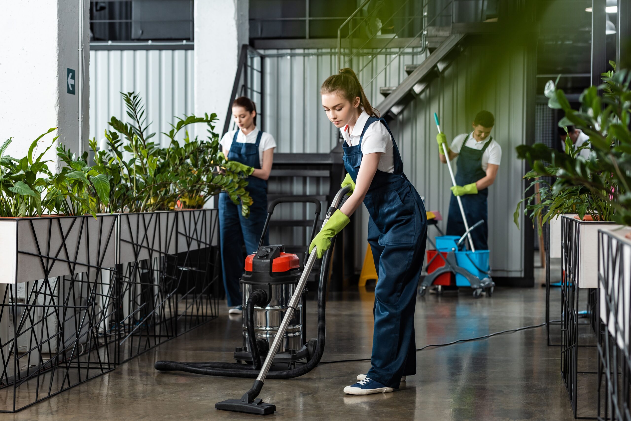Three workers in dark overalls clean a bright indoor space filled with plants. One uses a vacuum, another tends to plants, and a third mops.
