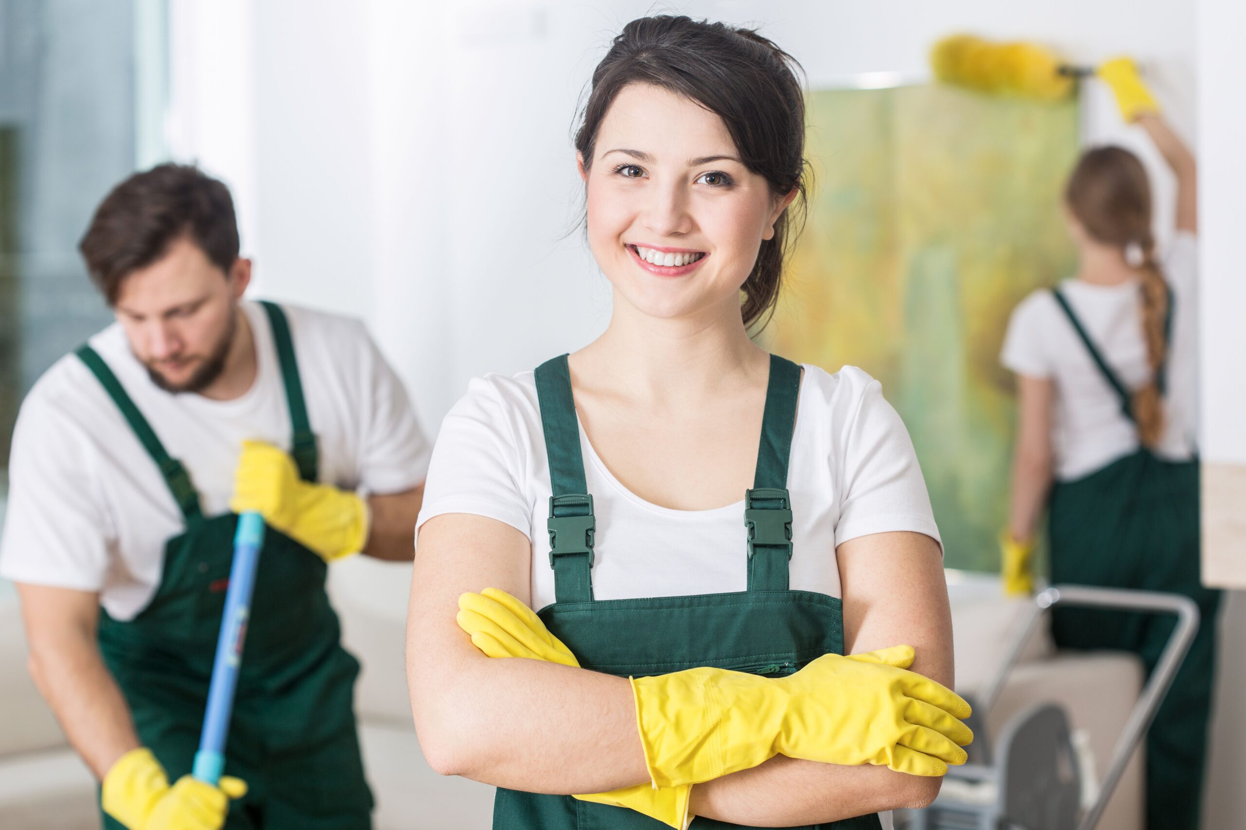 A smiling woman in green overalls and yellow gloves stands with her arms crossed, while colleagues clean the background. It's a team-focused cleaning setting.