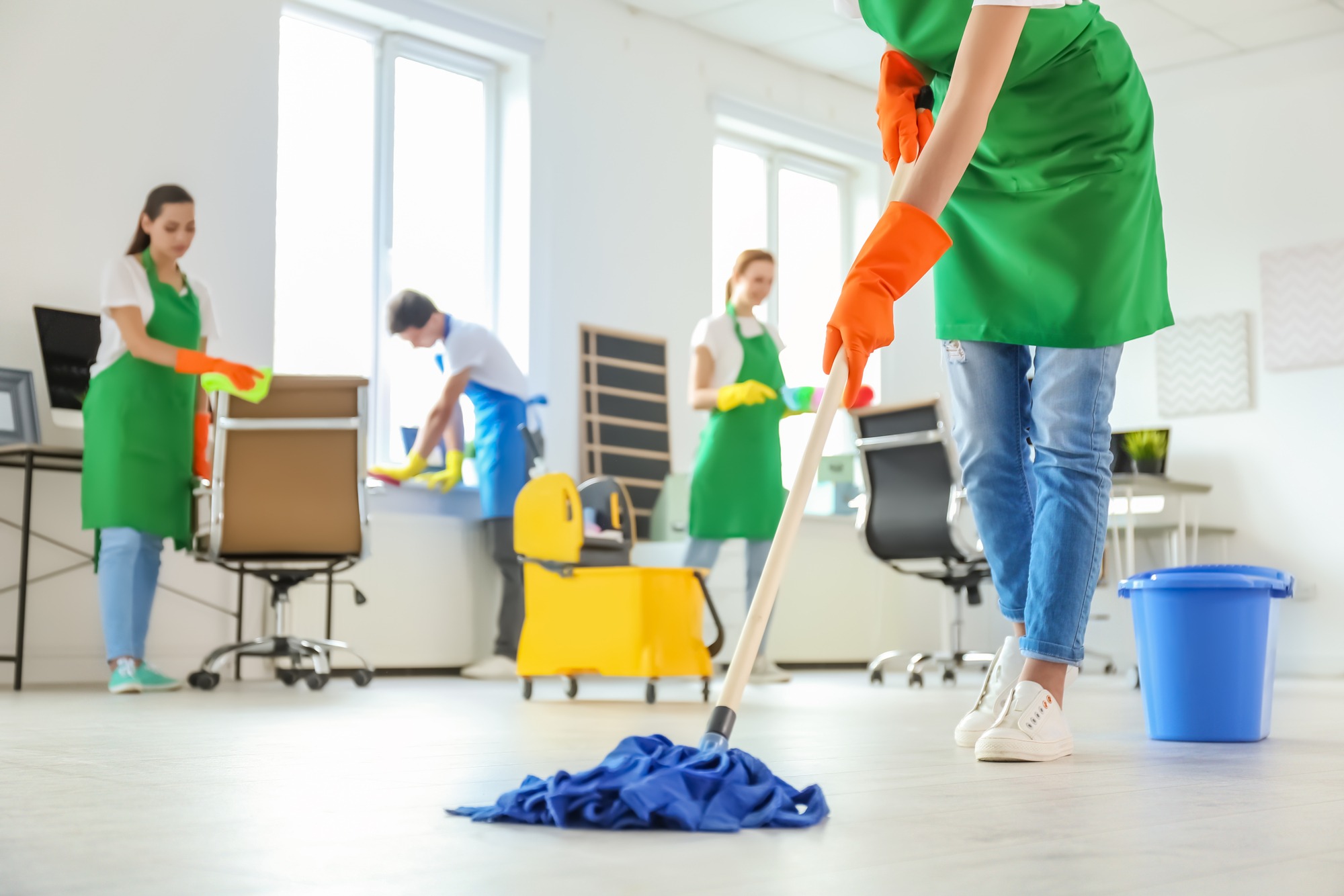 A group of workers in green aprons and gloves clean an office space, with one person mopping the floor while others wash windows and tidy desks.